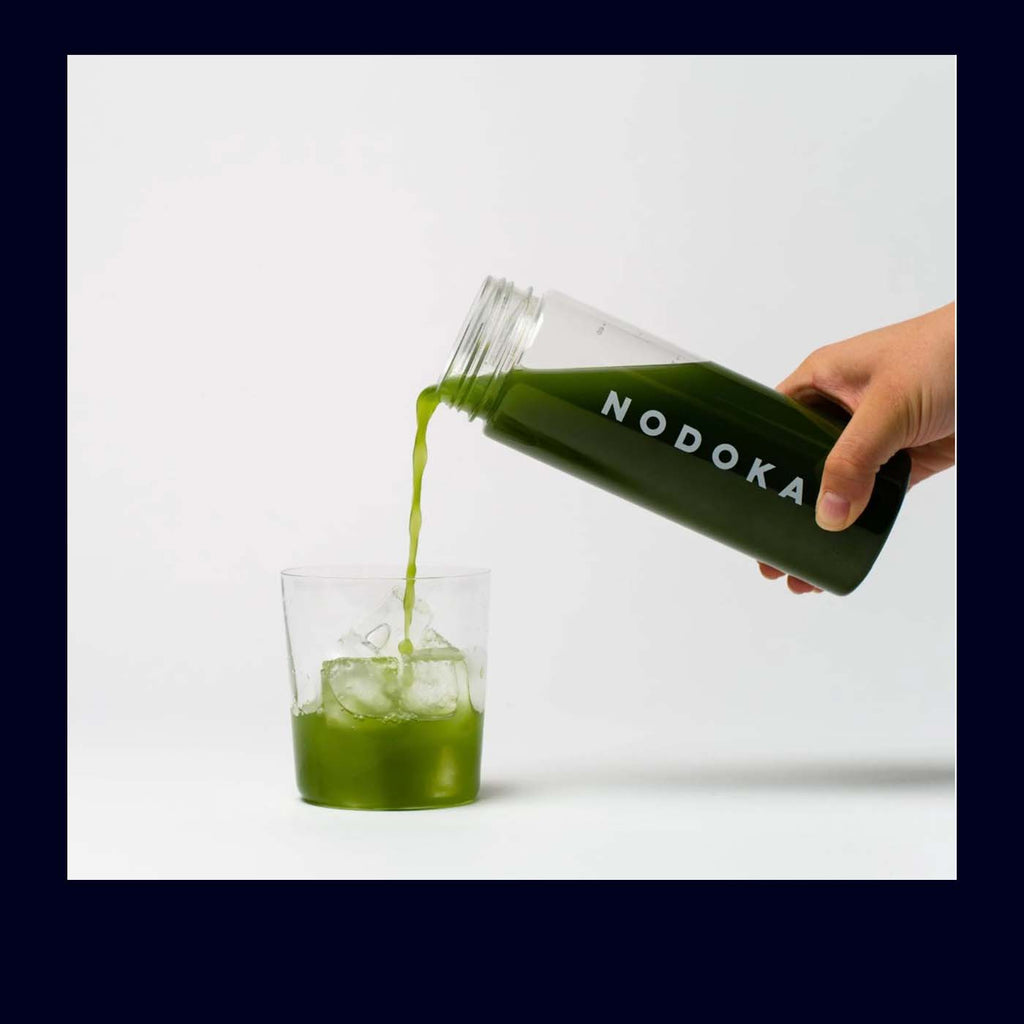 Hand pouring green matcha tea from a NODOKA bottle into a glass on a white background